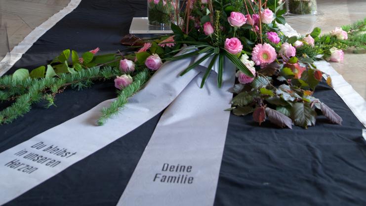 Blumenarrangement mit rosa Rosen und eine Trauerschleife vor einem Kreuz in einer Kapelle.