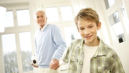 Ein Junge lächelt, während er die Hand eines älteren Mannes hält, in einem hellen Raum mit Fenstern.